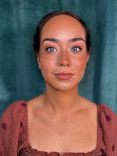 Load image into Gallery viewer, A close-up portrait of a woman with freckles and blue eyes, wearing a rust-colored textured top, set against a dark teal textured background.